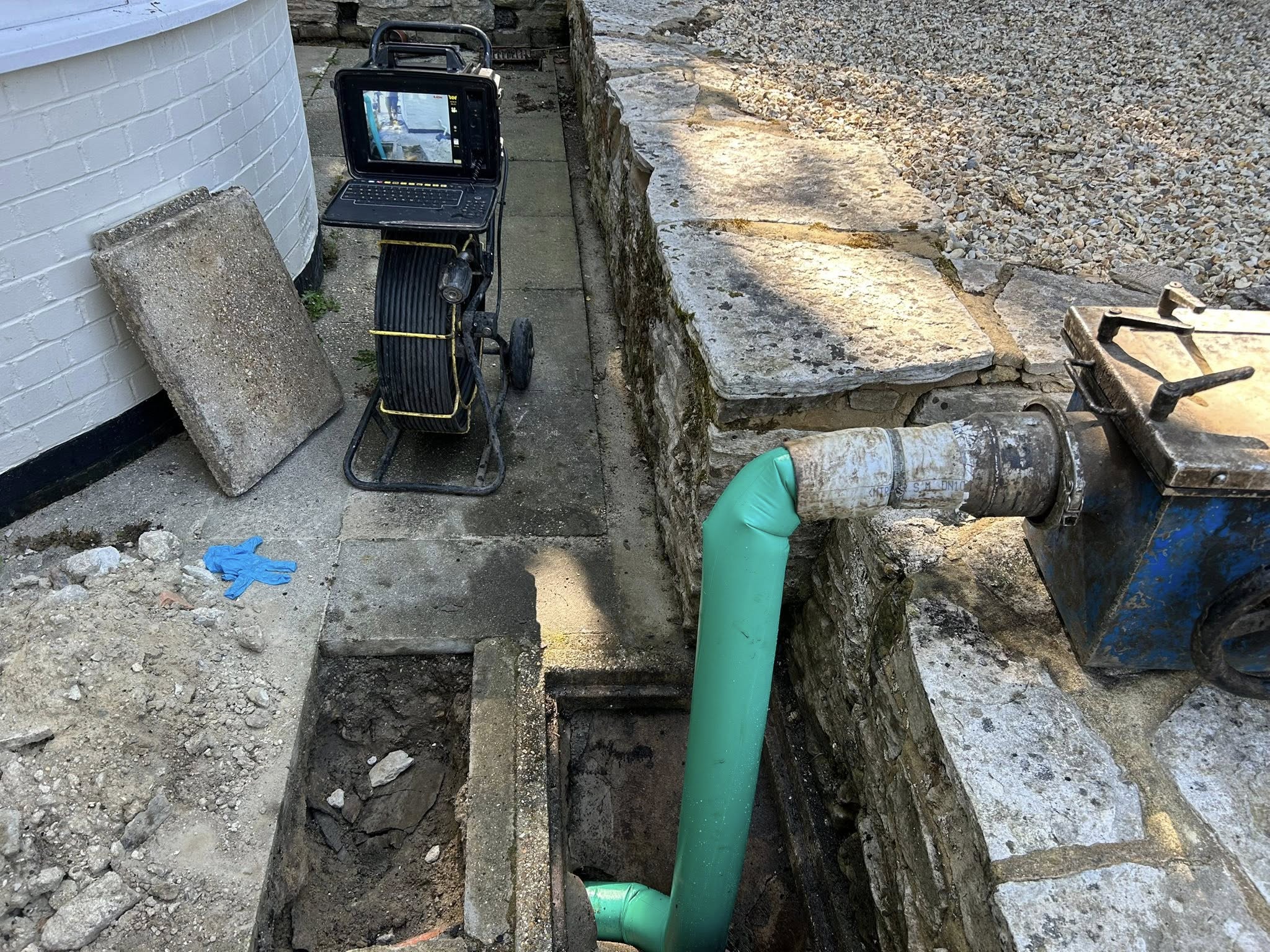 A green pipe connects to a large blue valve in a stone-walled trench. Nearby, a monitor is set up on a reel of cable, and a concrete slab rests on the paving stones beside the trench.