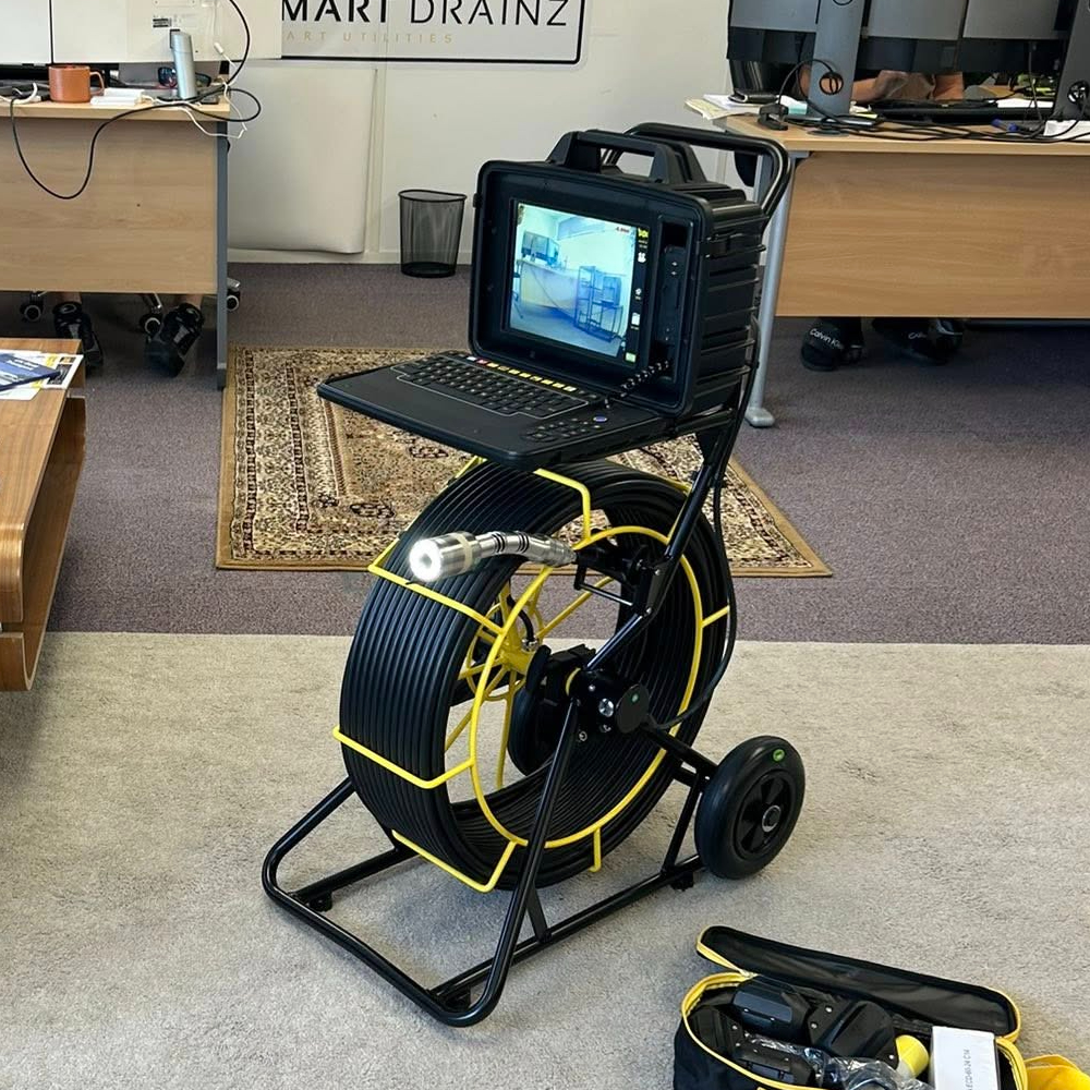 A wheeled inspection camera system with a monitor and long coiled cable is set up in an office, displaying a video feed. Desks, chairs, and a carpet are visible in the background.