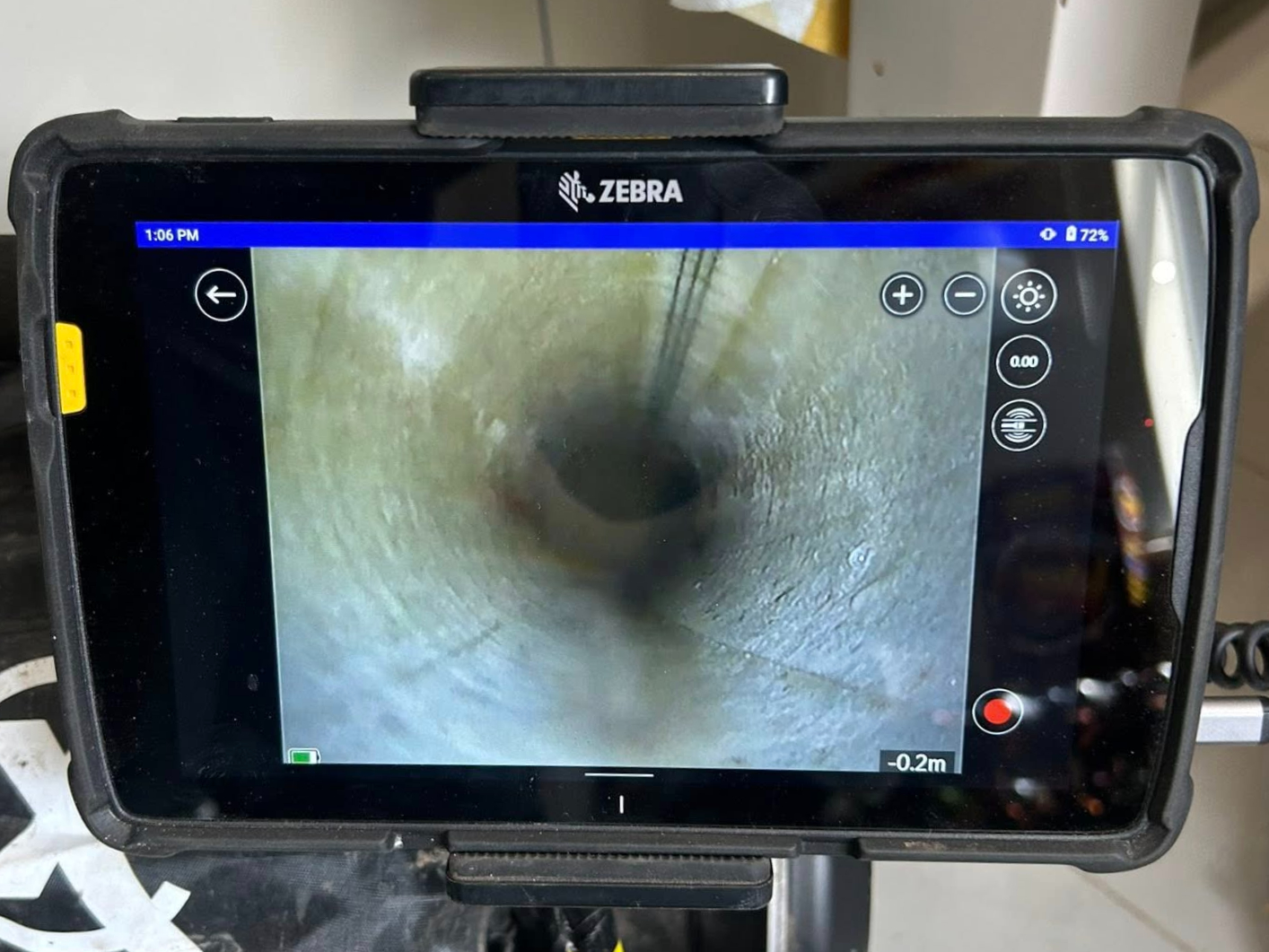 A tablet screen displays a live video feed from an inspection camera inside a pipe, showing the pipe’s inner surface and a cable or tool partially visible in the center. On-screen controls and status icons are visible.