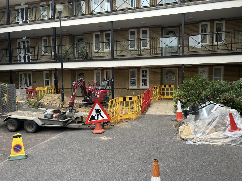 Construction work outside an apartment building with barriers, cones, a small excavator, a trailer, and a wheelbarrow. A "men at work" sign is in front, with some soil and debris on the ground.