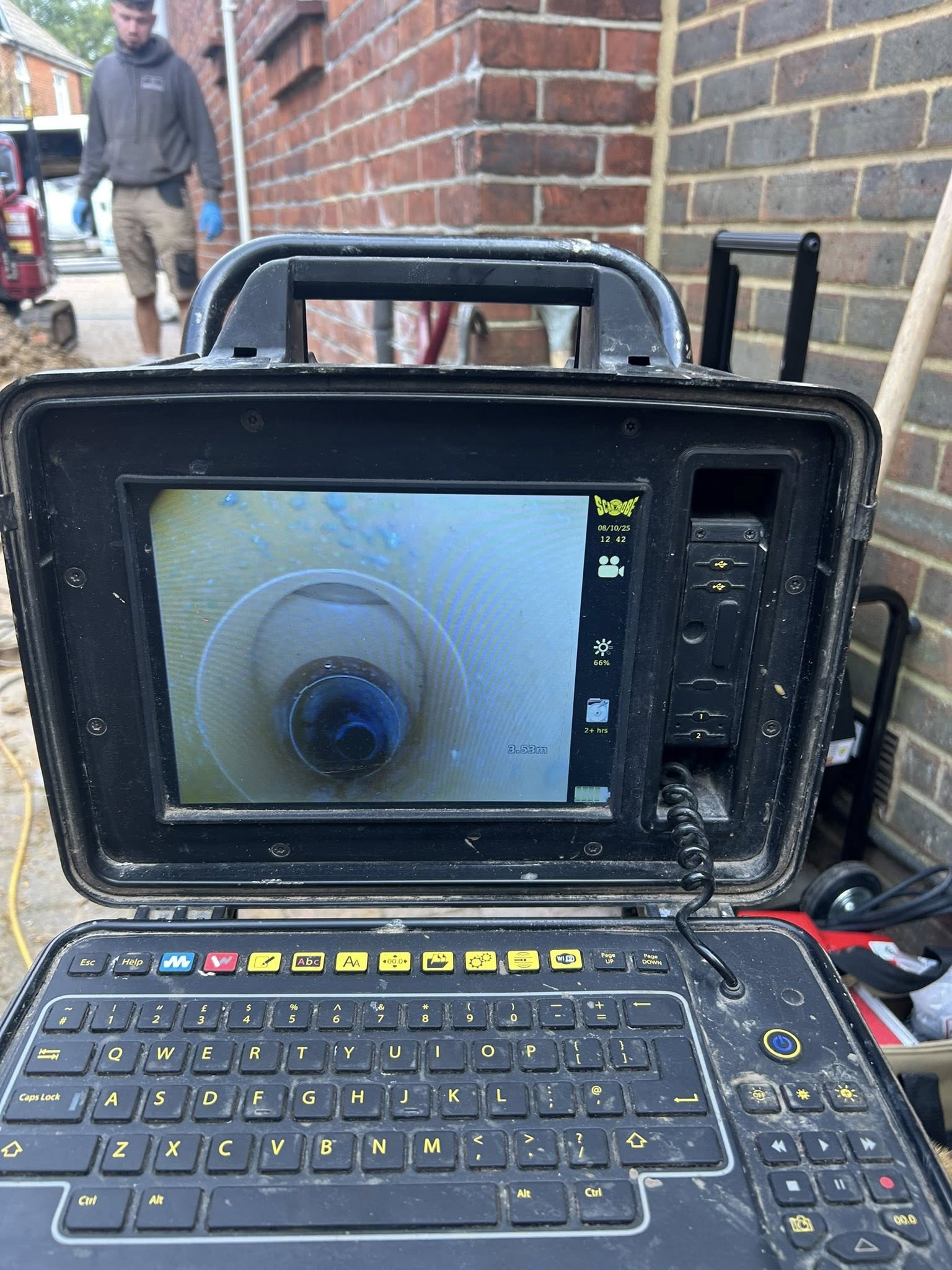 A close-up of a rugged inspection camera monitor showing the inside of a pipe. The device sits on the ground near a brick wall, with a man standing in the background. Various cables and tools are visible around.