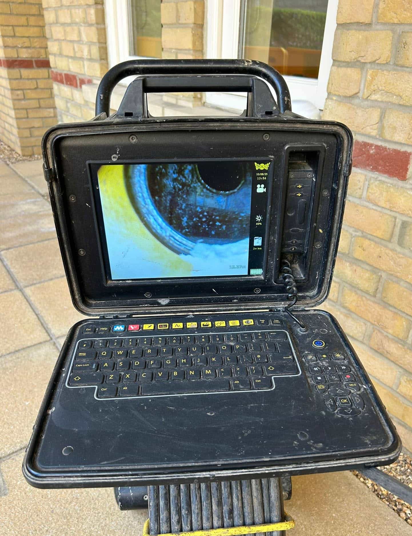 A rugged, black, portable inspection computer with a built-in keyboard displays a close-up image of a pipe or tunnel interior. The device is outdoors on a paved surface near a brick wall and window.