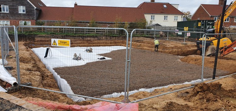 A fenced construction site with a gravel-filled rectangular foundation, bordered by white sheeting. A warning sign and two workers in safety gear are present. Houses and construction equipment are visible in the background.