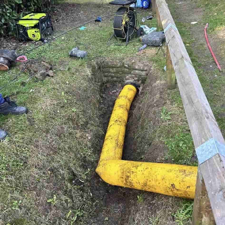 A large yellow pipe runs underground in a dug trench next to a wooden fence, with tools and equipment scattered nearby on the grass.
