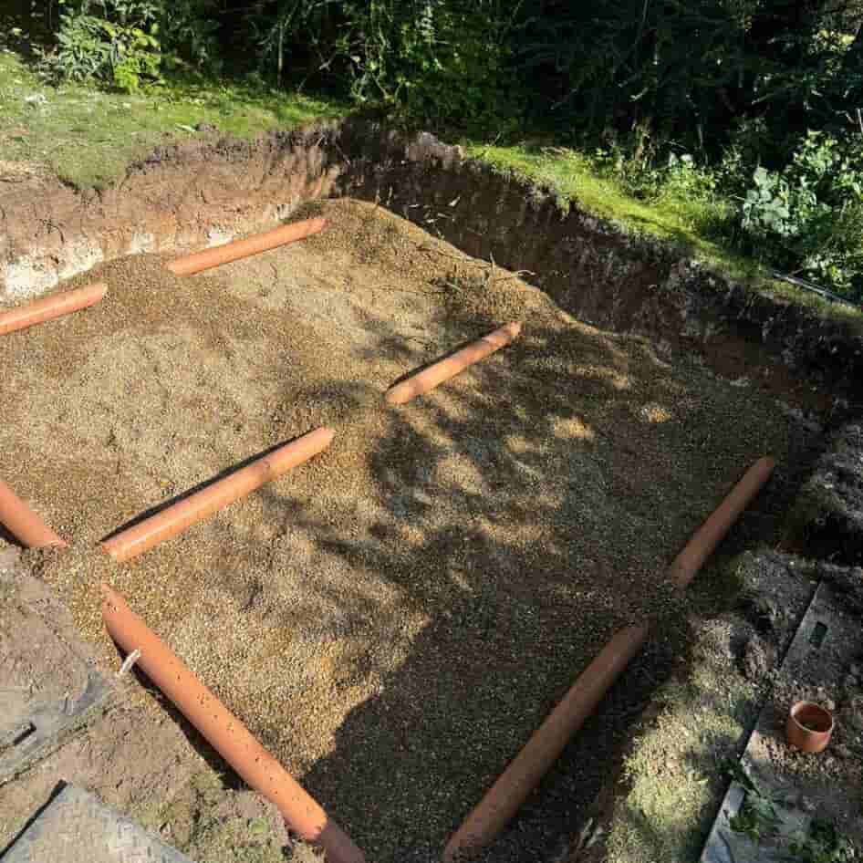 A rectangular pit in the ground filled with gravel and five evenly spaced horizontal orange drainage pipes, surrounded by grass and vegetation.