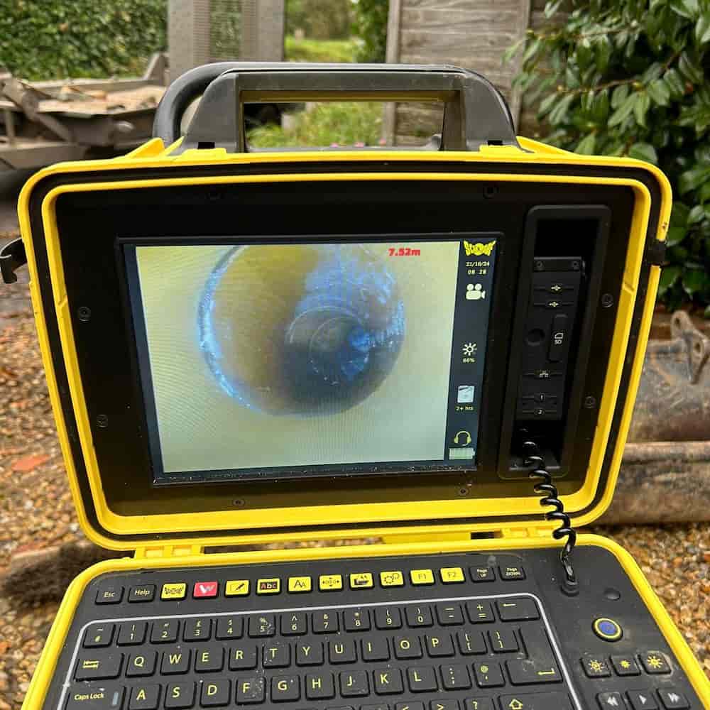 A rugged, yellow inspection camera displays an image of a pipe’s interior on its screen, showing a round, dark obstruction. The device has a keyboard and controls below the screen.