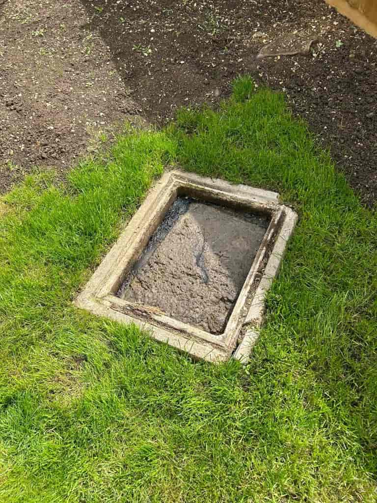 A muddy, water-filled rectangular drain or inspection chamber is set in the ground, surrounded by green grass and soil.