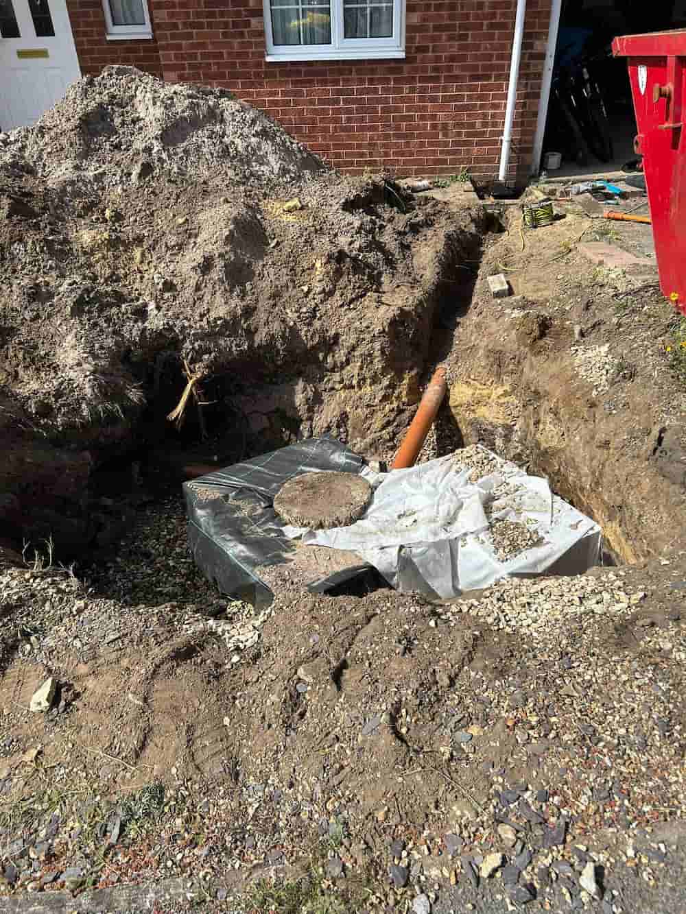 A large hole dug in front of a brick house reveals a partially buried septic tank with a pipe attached, surrounded by dirt, gravel, and construction materials.