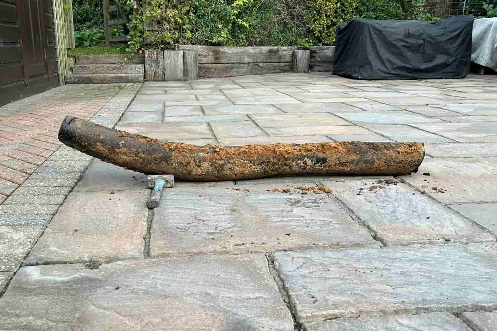 A large, rusty, and bent metal pipe lies on a stone patio, with a small wrench attached to one end. There are garden beds and greenery in the background.