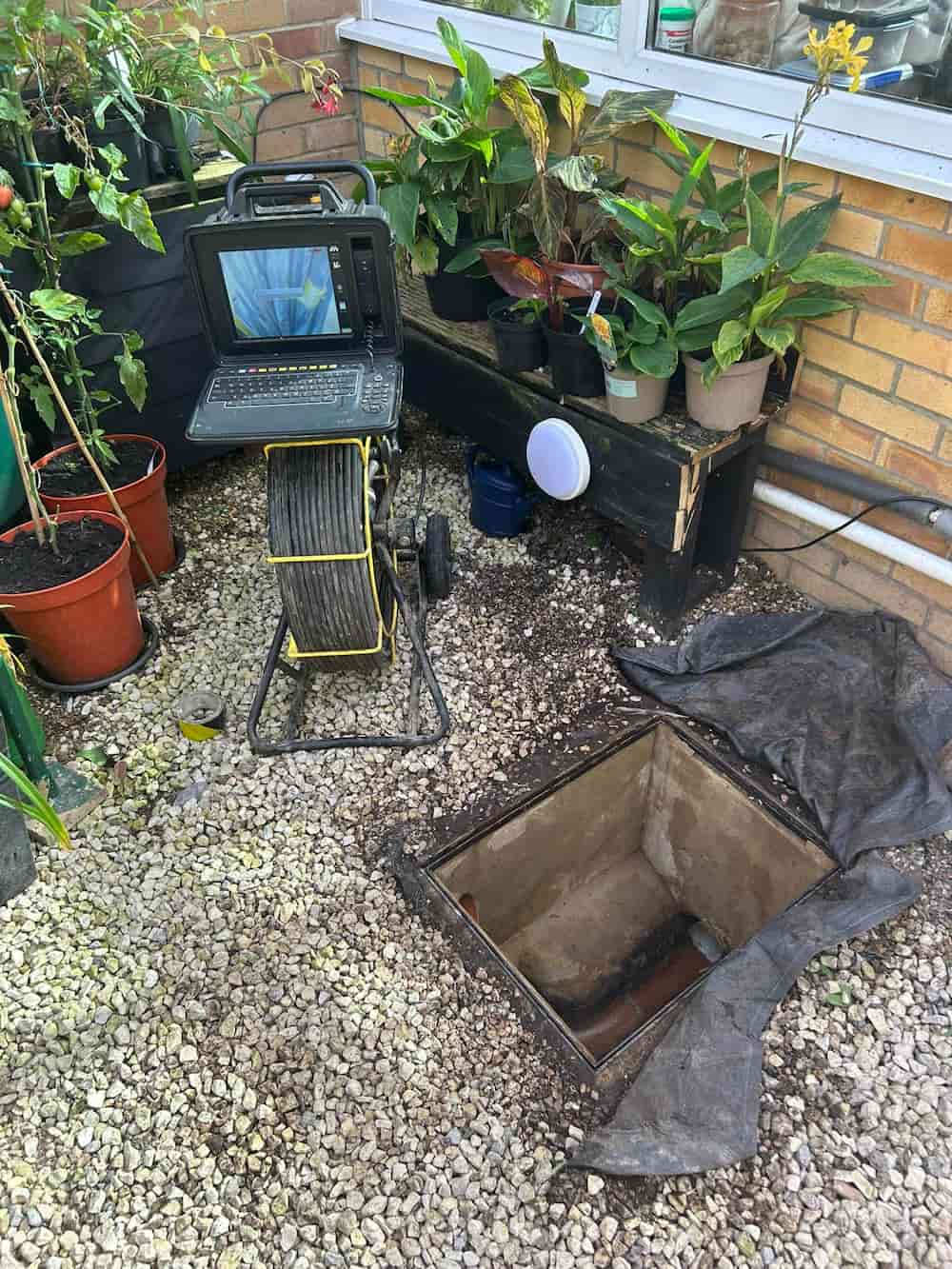 A drain inspection camera with monitor is set up beside an open inspection chamber surrounded by potted plants and gravel in a garden area next to a house wall.