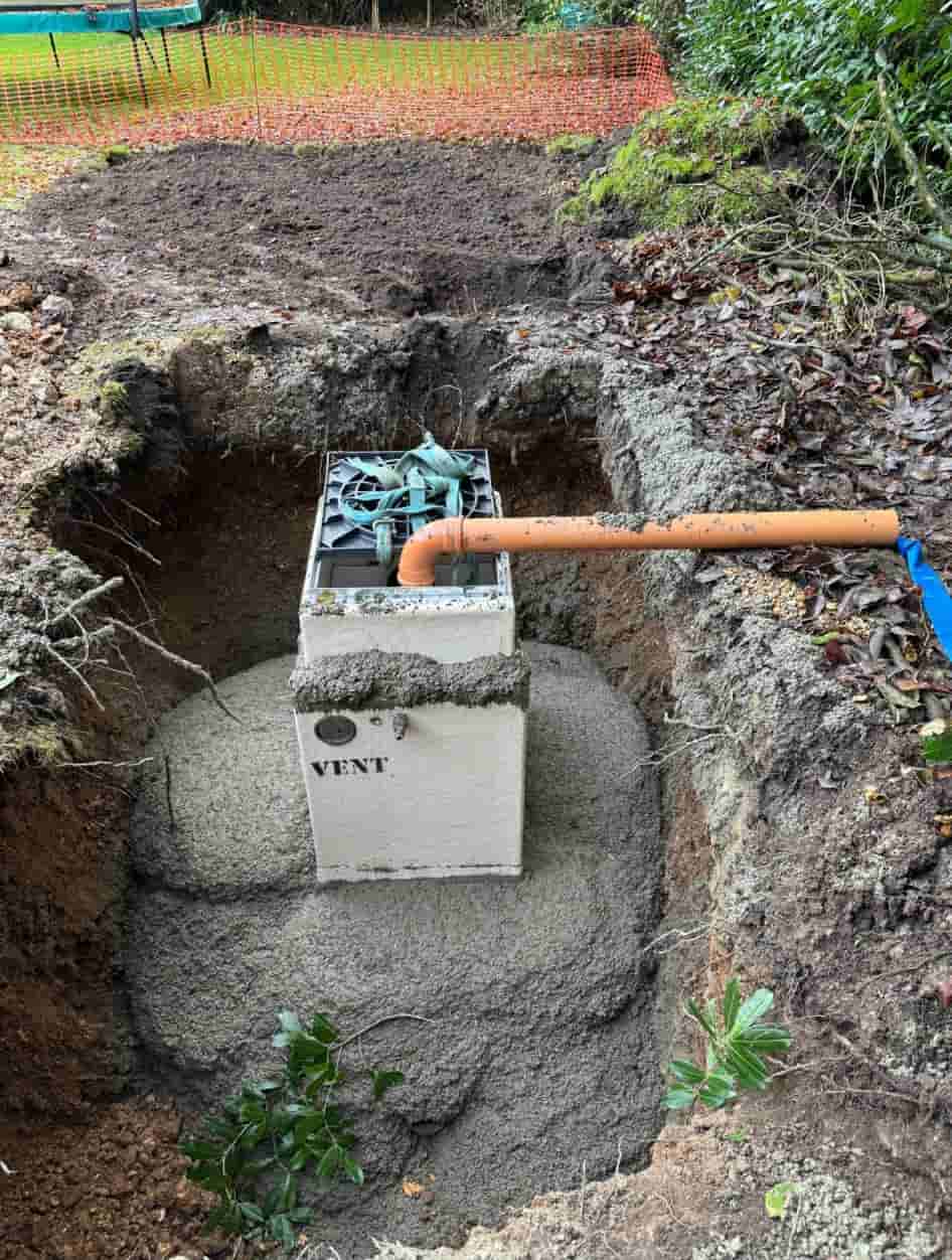 A white septic tank partially buried in a dirt trench, with a vent and an orange pipe connected. The tank is surrounded by soil and vegetation, with construction equipment and safety fencing visible in the background.