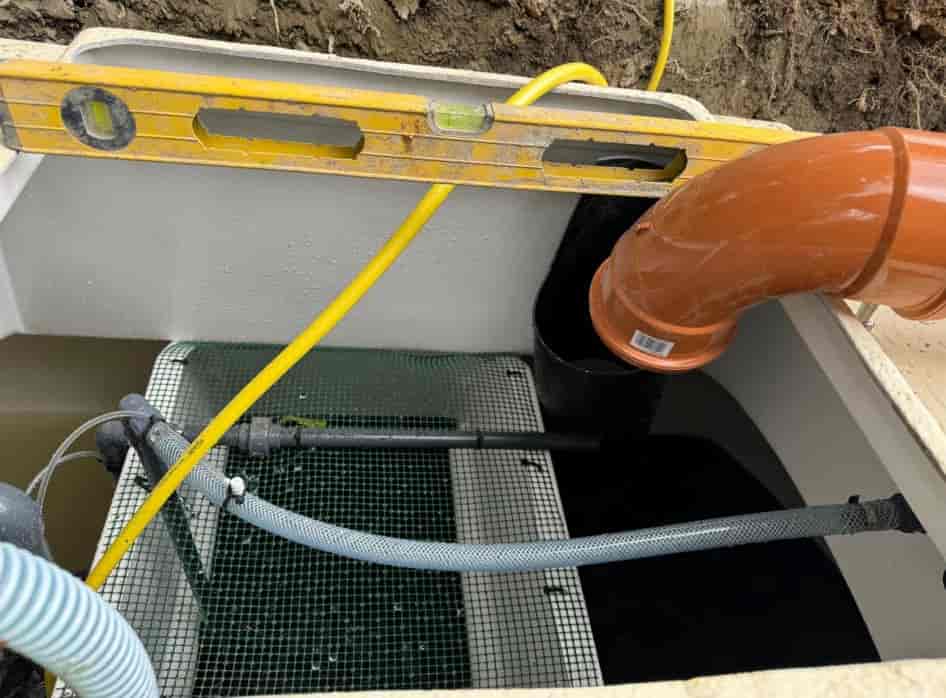 A close-up view of a water filtration or drainage system, featuring a mesh-covered compartment, pipes, an orange elbow joint, a yellow hose, and a level tool resting across the top.