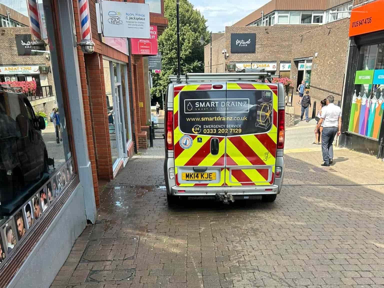 A white van with red and yellow chevron stripes and "Smart Drainz" branding is parked on a pedestrian walkway next to shops. People walk nearby in a shopping area on a sunny day.