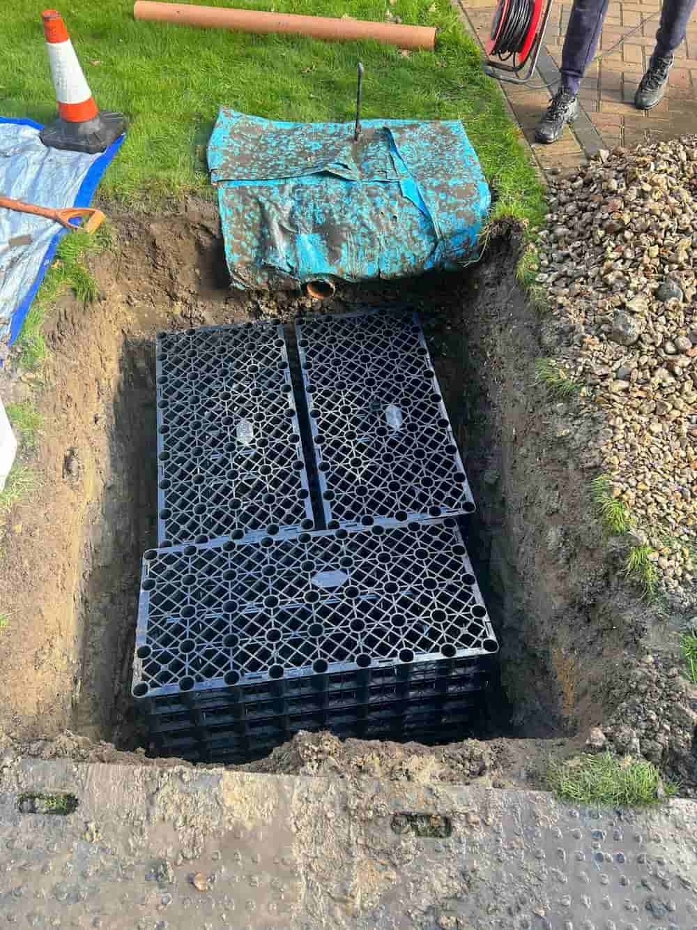 Two large black plastic soakaway crates installed in a rectangular hole dug in a grassy area, with tools, a blue tarp, and a traffic cone nearby. Only a person's lower legs and feet are visible at the edge.