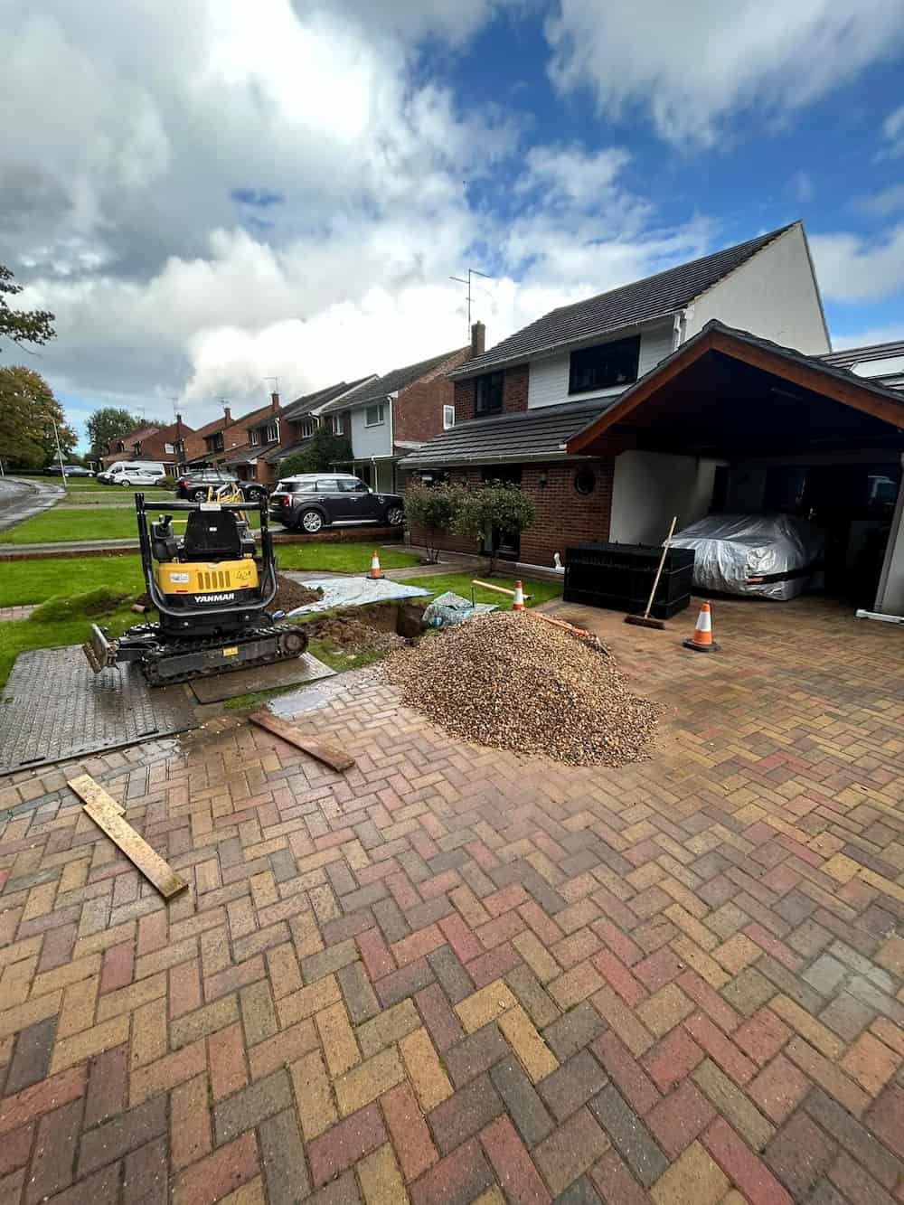 A paved driveway in front of a suburban house, with a small excavator, a pile of gravel, traffic cones, and some wooden planks, indicating ongoing construction or landscaping work.