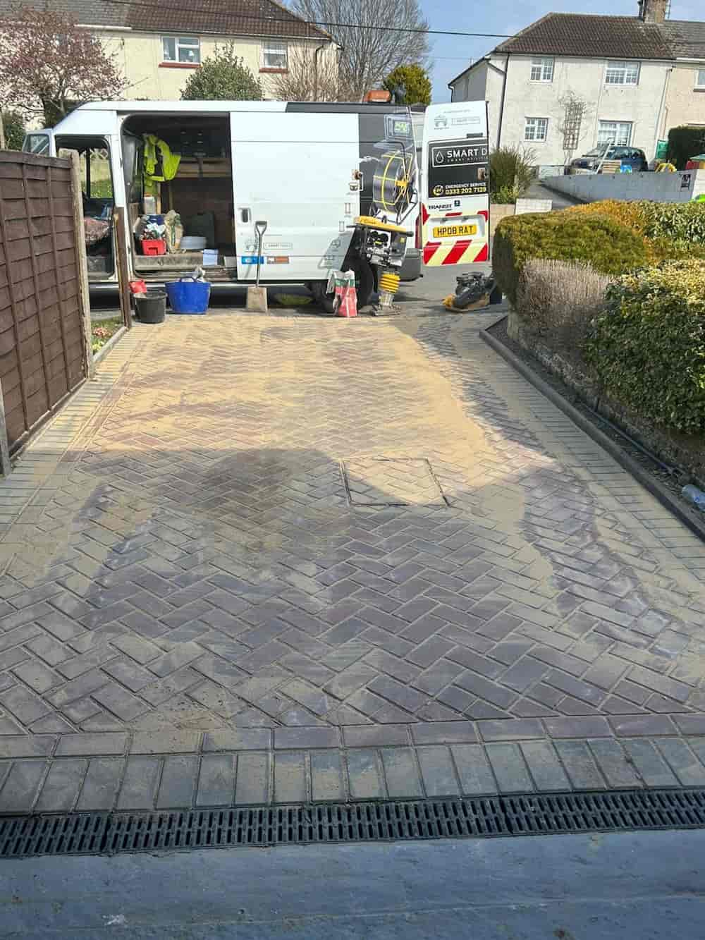 A newly laid herringbone-pattern driveway bordered by hedges and a wooden fence, with a work van and equipment parked at the end, and sand spread over the bricks.