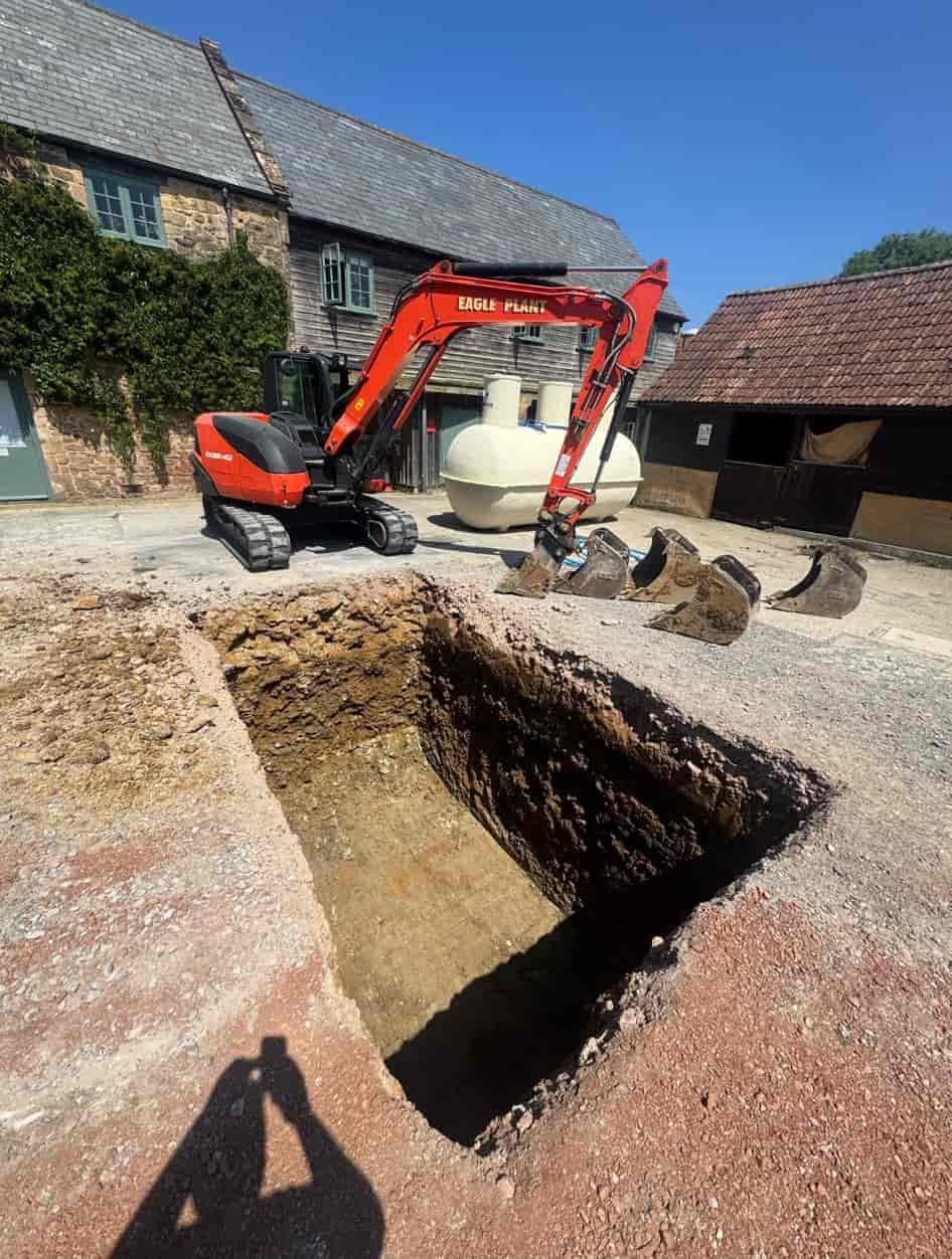A red excavator is parked beside a large, freshly dug rectangular pit on a gravel surface near old stone buildings with tiled roofs.