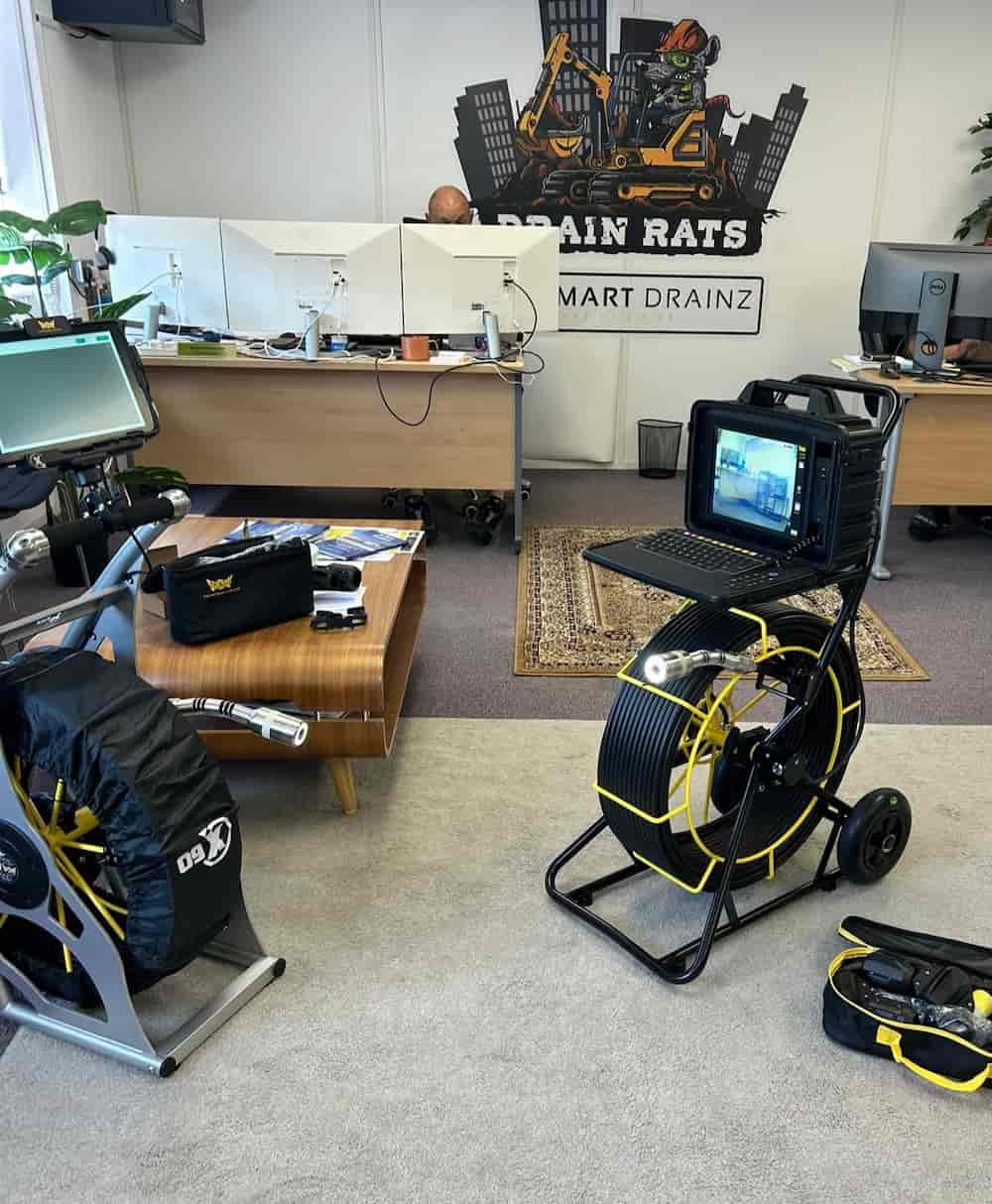 An office with desks, computers, and a large wall logo reading "Drain Rats Smart Drainz." In the foreground is a drain inspection camera system on wheels with cables and a monitor showing footage.