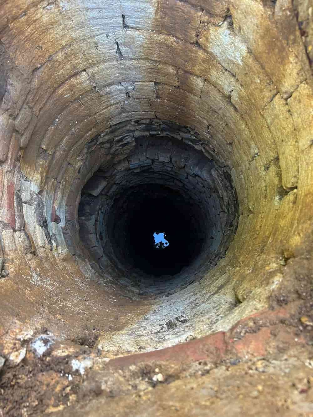 View looking down into a deep, circular stone well, with light visible at the bottom and rough, weathered bricks lining the walls.