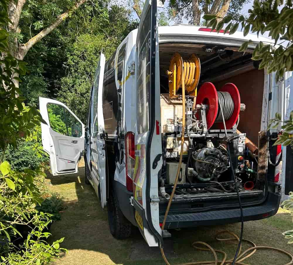 A white service van with its back and side doors open is parked on grass. Inside the van are large hoses on reels and cleaning equipment, surrounded by trees and greenery.