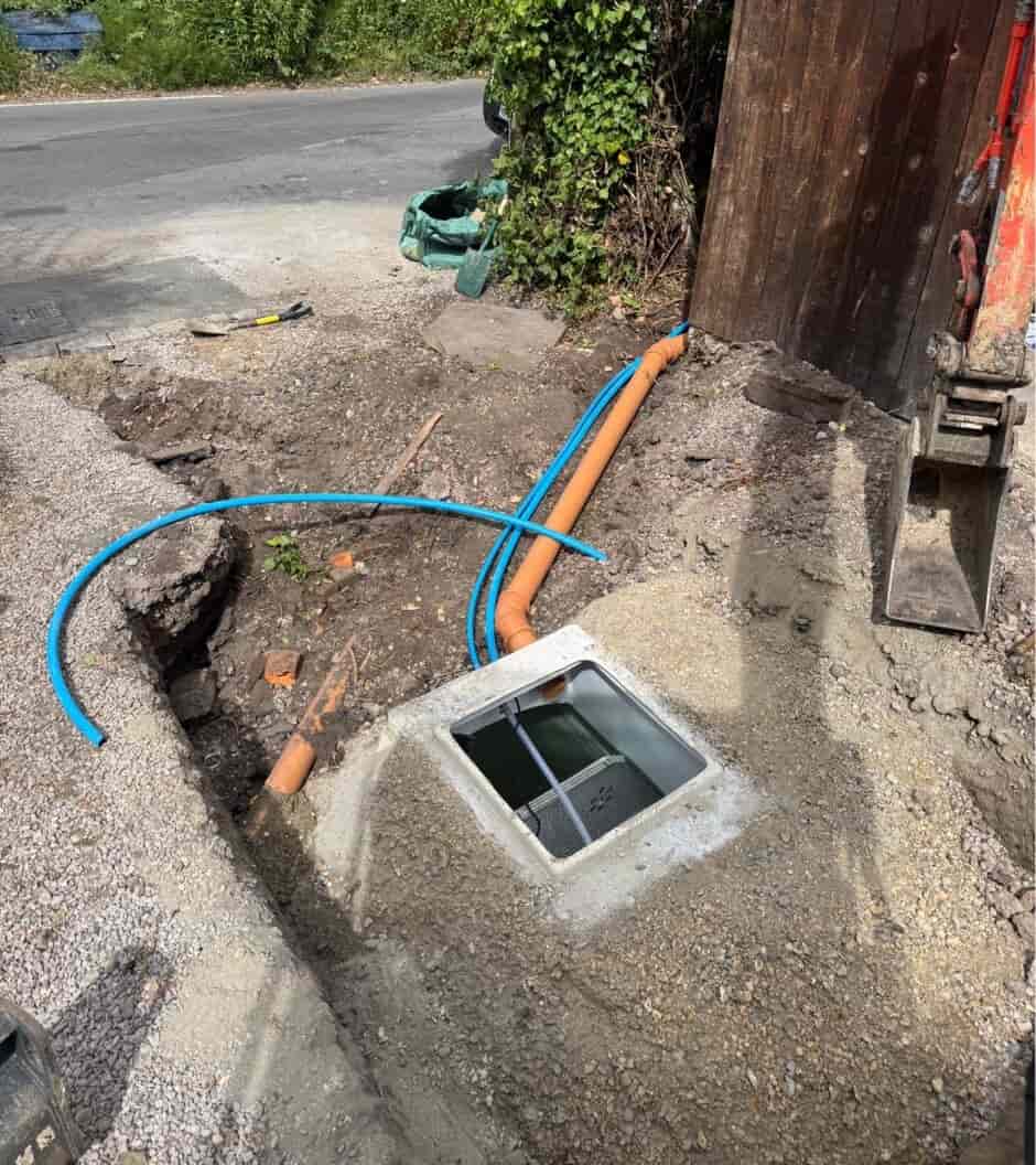A construction site with an open drainage or utility box set in gravel, blue and orange pipes leading into it, and a partially dug trench beside a road with green foliage in the background.