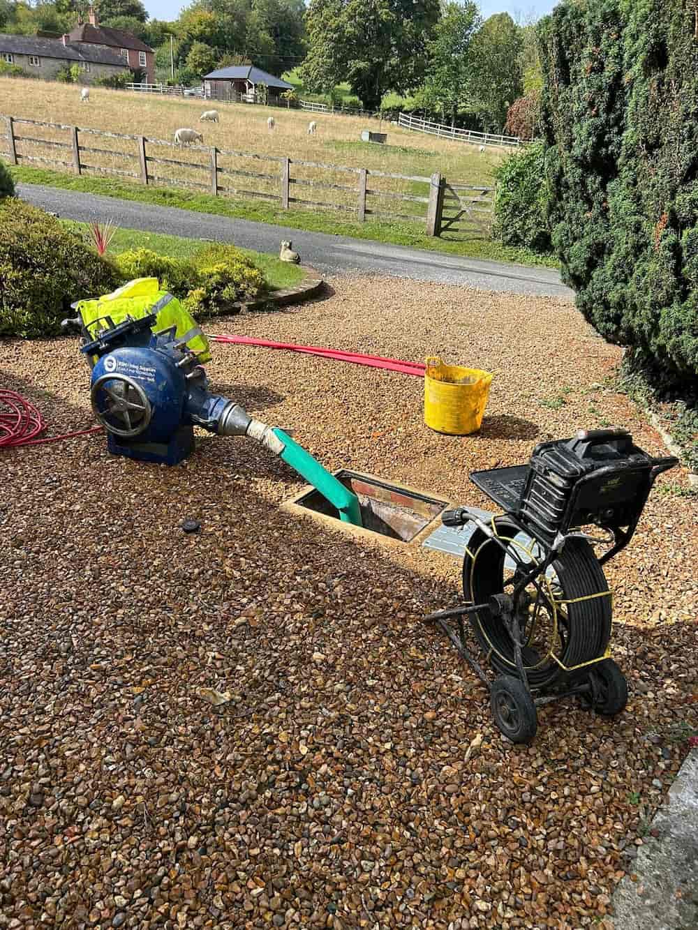 A mechanical pump and a cable reel are set up on a gravel driveway, with a green hose going into an open manhole. There’s a yellow bucket nearby, and sheep can be seen in a field in the background.