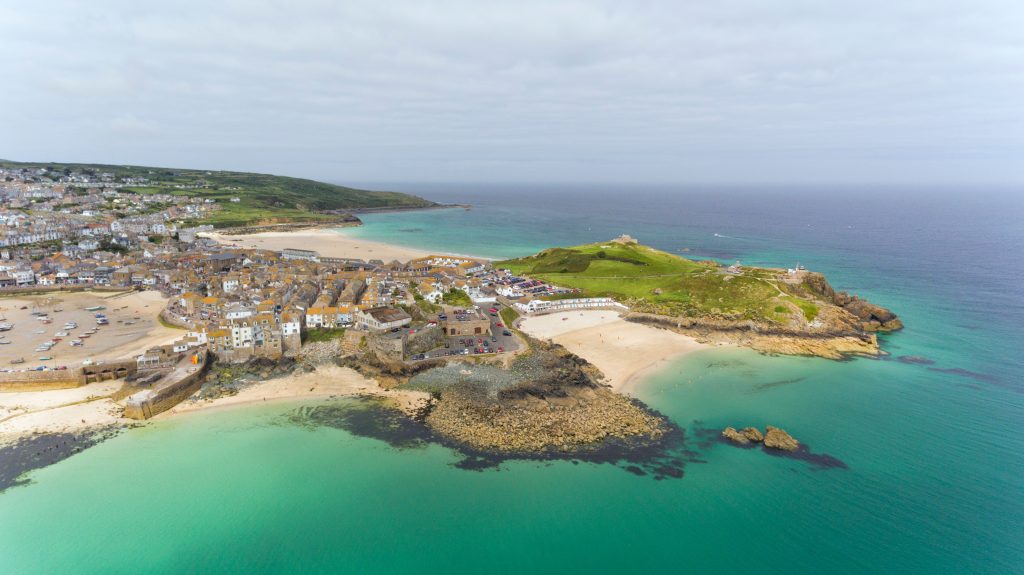Aerial view of a coastal town with sandy beaches, turquoise sea, rocky shorelines, clustered buildings, and green hills under a cloudy sky.