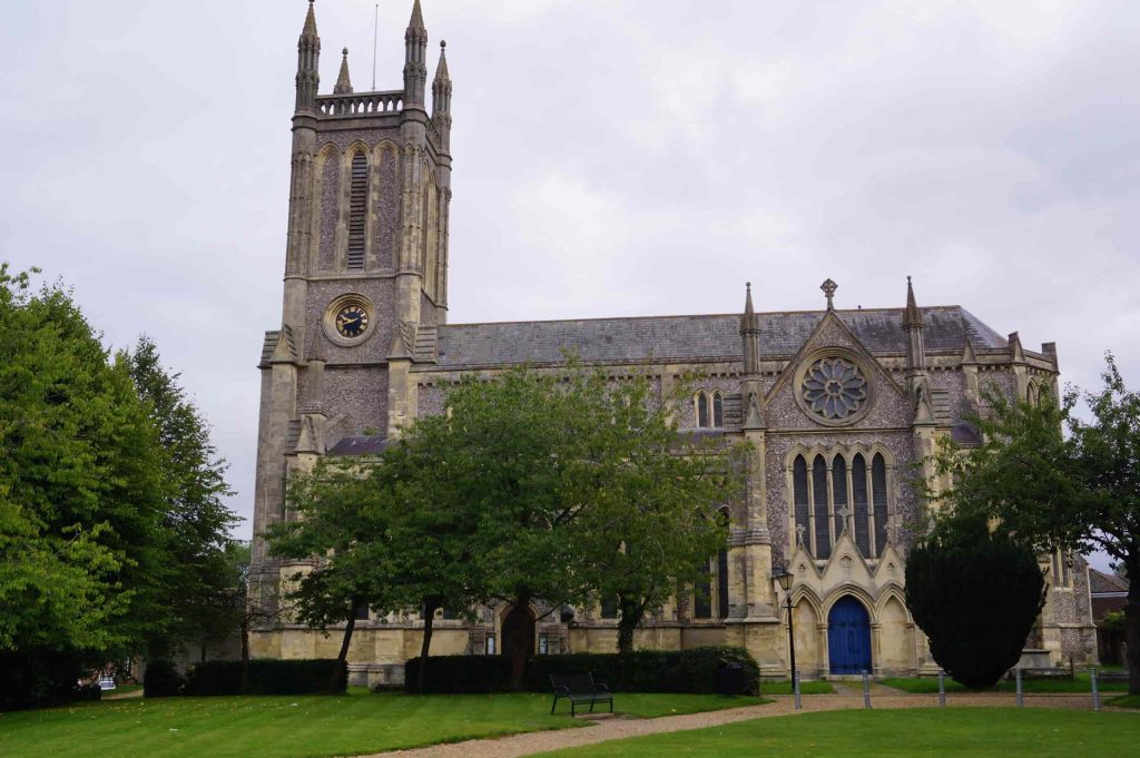 A large stone church with a tall clock tower, pointed arches, stained glass windows, and blue doors, surrounded by green trees and grass under a cloudy sky.