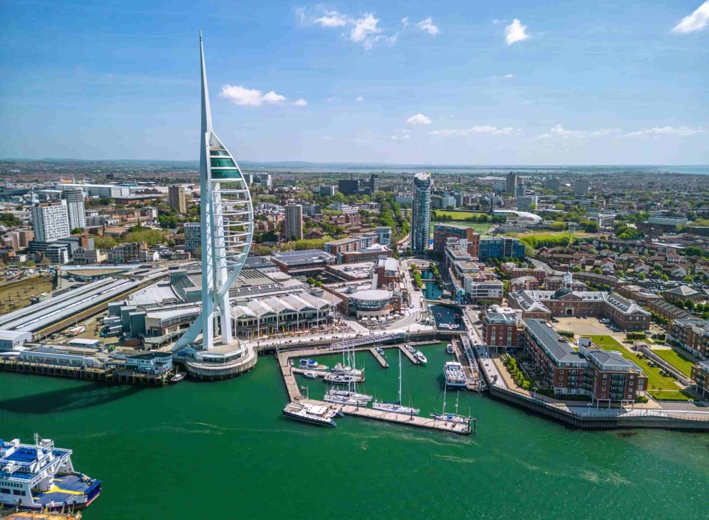 Aerial view of Portsmouth, UK, featuring the white Spinnaker Tower near the waterfront, surrounded by boats, docks, modern buildings, and cityscape under a blue sky.