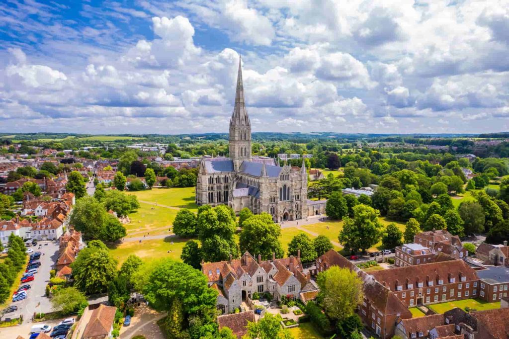 Aerial view of Salisbury Cathedral with its tall spire, surrounded by green lawns, trees, and historic buildings under a partly cloudy sky in Salisbury, England.