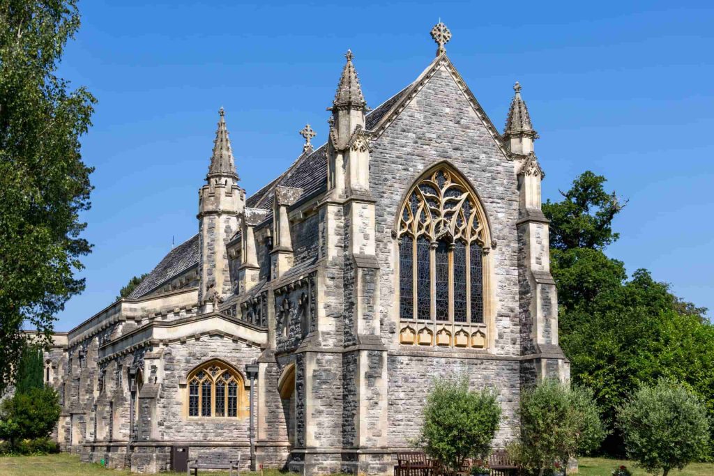 A stone Gothic-style church with pointed arches, large stained-glass windows, and twin spires, surrounded by green trees and shrubs under a clear blue sky.
