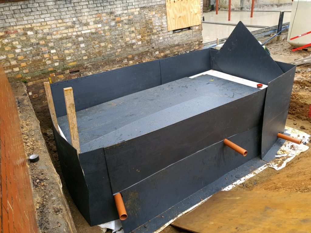 A large, rectangular, black waterproof tank with two orange pipes protruding from the sides sits in an outdoor construction site near a brick wall. The tank is surrounded by soil and construction materials.