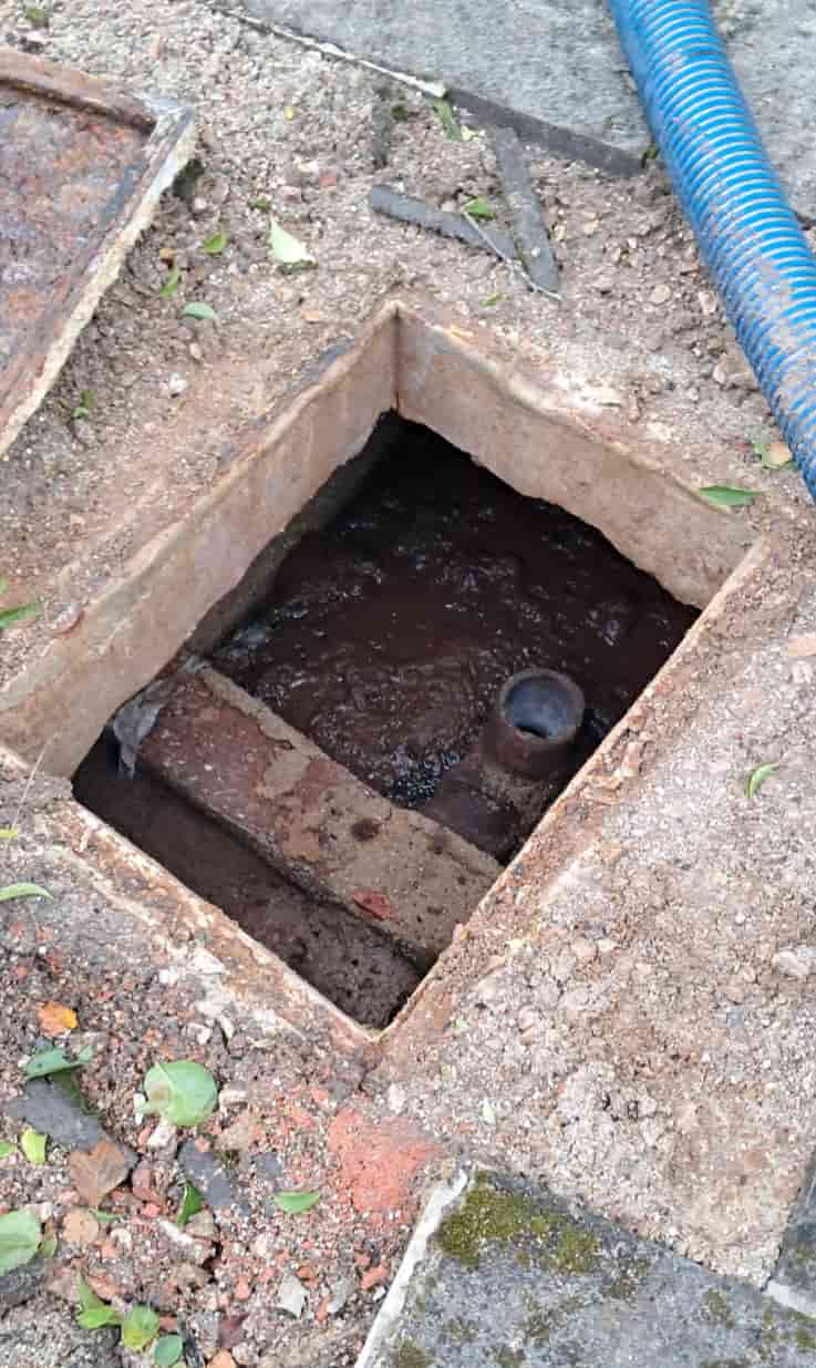 A rectangular open manhole filled with dark, muddy water and debris. A blue flexible hose is positioned nearby, and there are leaves and dirt around the opening.