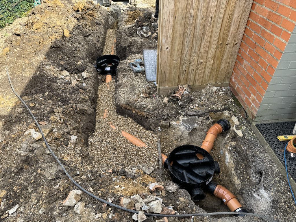 A trench dug alongside a brick building contains new underground drainage pipes and inspection chambers, surrounded by gravel and soil, with construction tools and debris nearby.