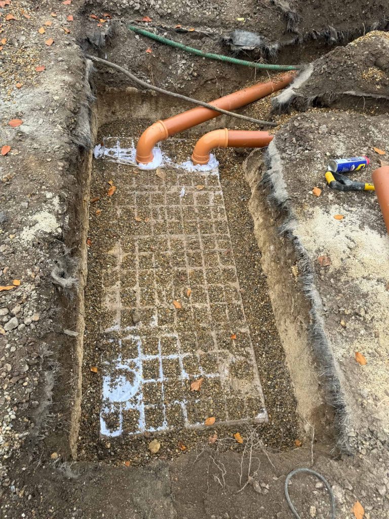 A gravel trench with two orange drainage pipes leading into it, partially filled and surrounded by soil. A can of construction adhesive and a tool are visible on the right edge of the trench.