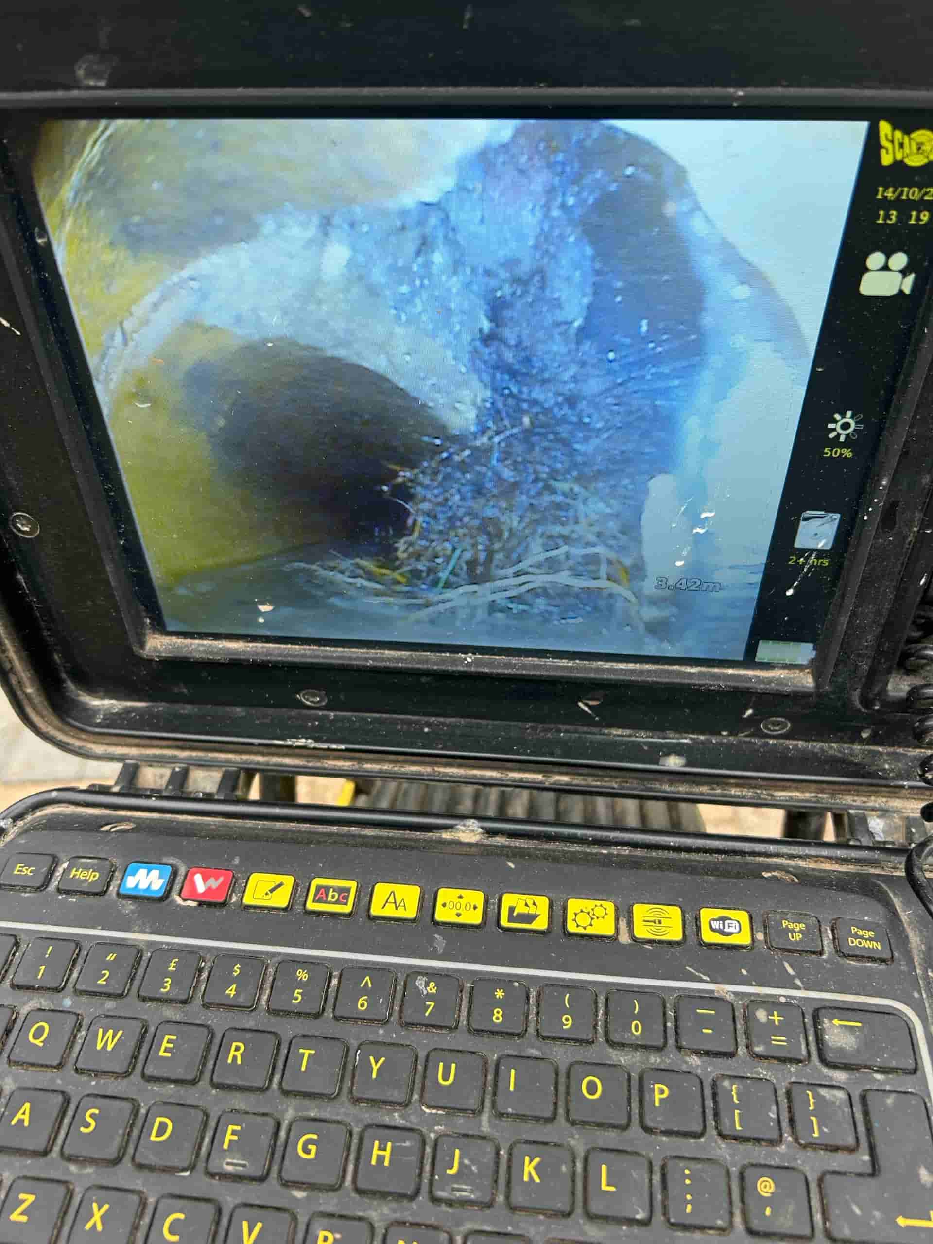 A monitor displays a close-up camera view inside a pipe, showing a blockage of roots. Below the screen is a keyboard with brightly colored keys, suggesting the use of a sewer inspection camera system.