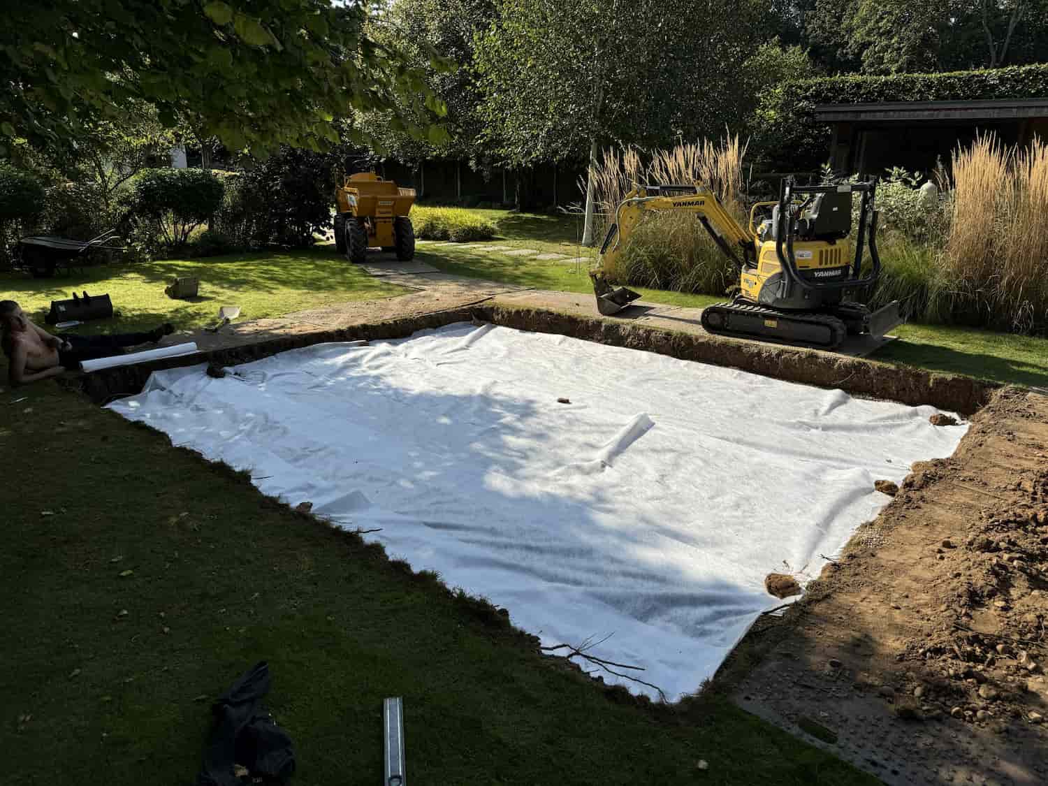 A rectangular area in a garden has been excavated, revealing white ground fabric. A yellow mini excavator and a yellow dump truck are nearby, surrounded by trees and tall grasses in the background.