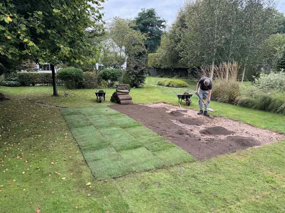 A person is laying turf in a garden, with several rolls of turf and patches of new grass on the ground. They are spreading soil with a shovel, surrounded by trees and shrubs on a cloudy day.