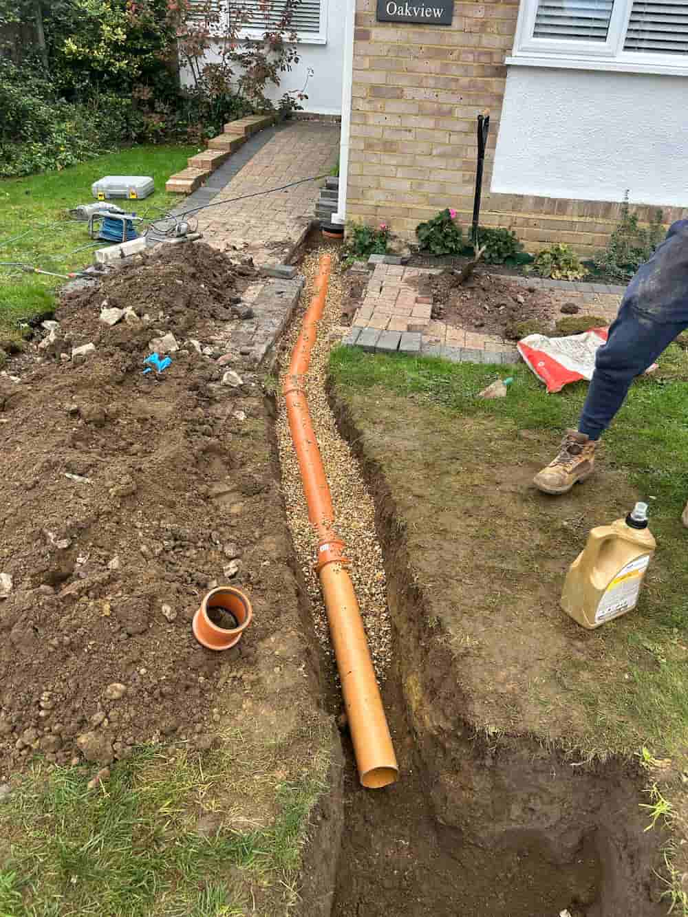 A trench with an orange drainage pipe being installed alongside a house, surrounded by dirt piles, gravel, tools, and construction materials. A person stands nearby on the right.