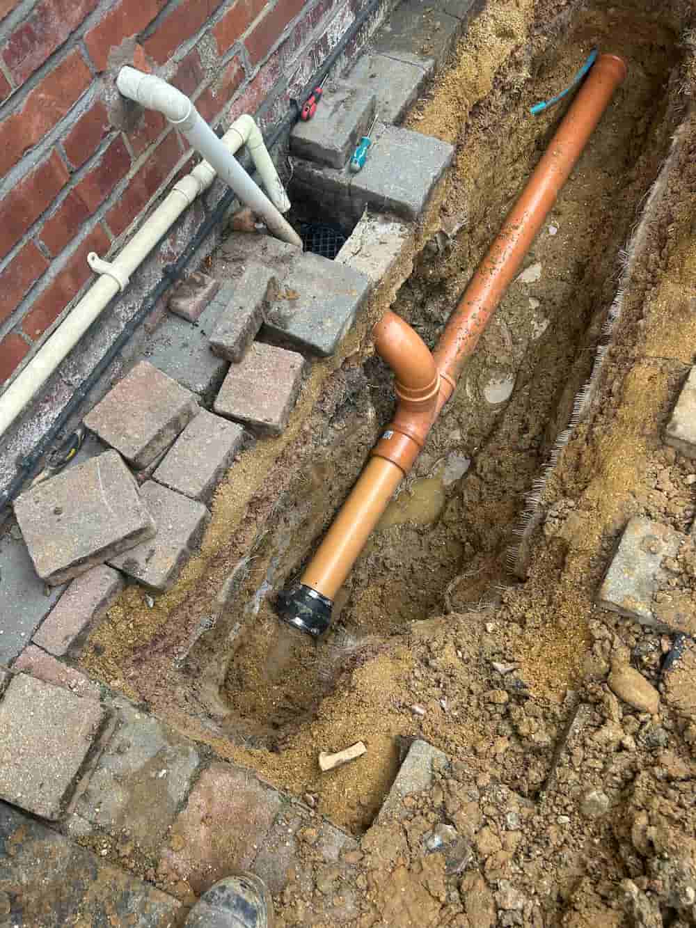An orange drainage pipe is being installed in a trench beside a brick wall. Paving stones have been lifted and are stacked nearby, and tools are visible around the work area.