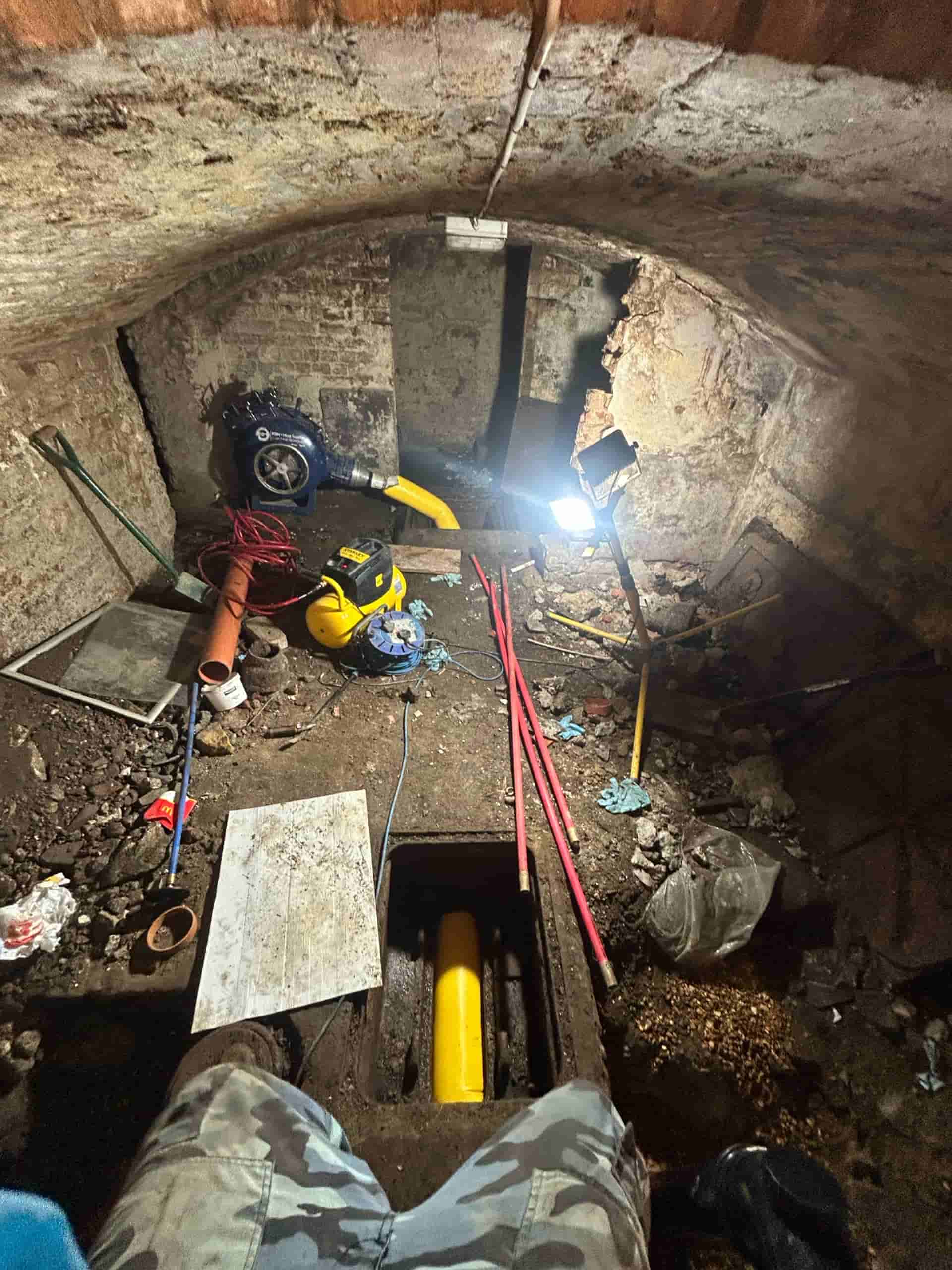 Dimly lit basement with arched brick ceiling, scattered construction tools, pipes, debris, and a bright work light illuminating the rough stone walls and cluttered floor.
