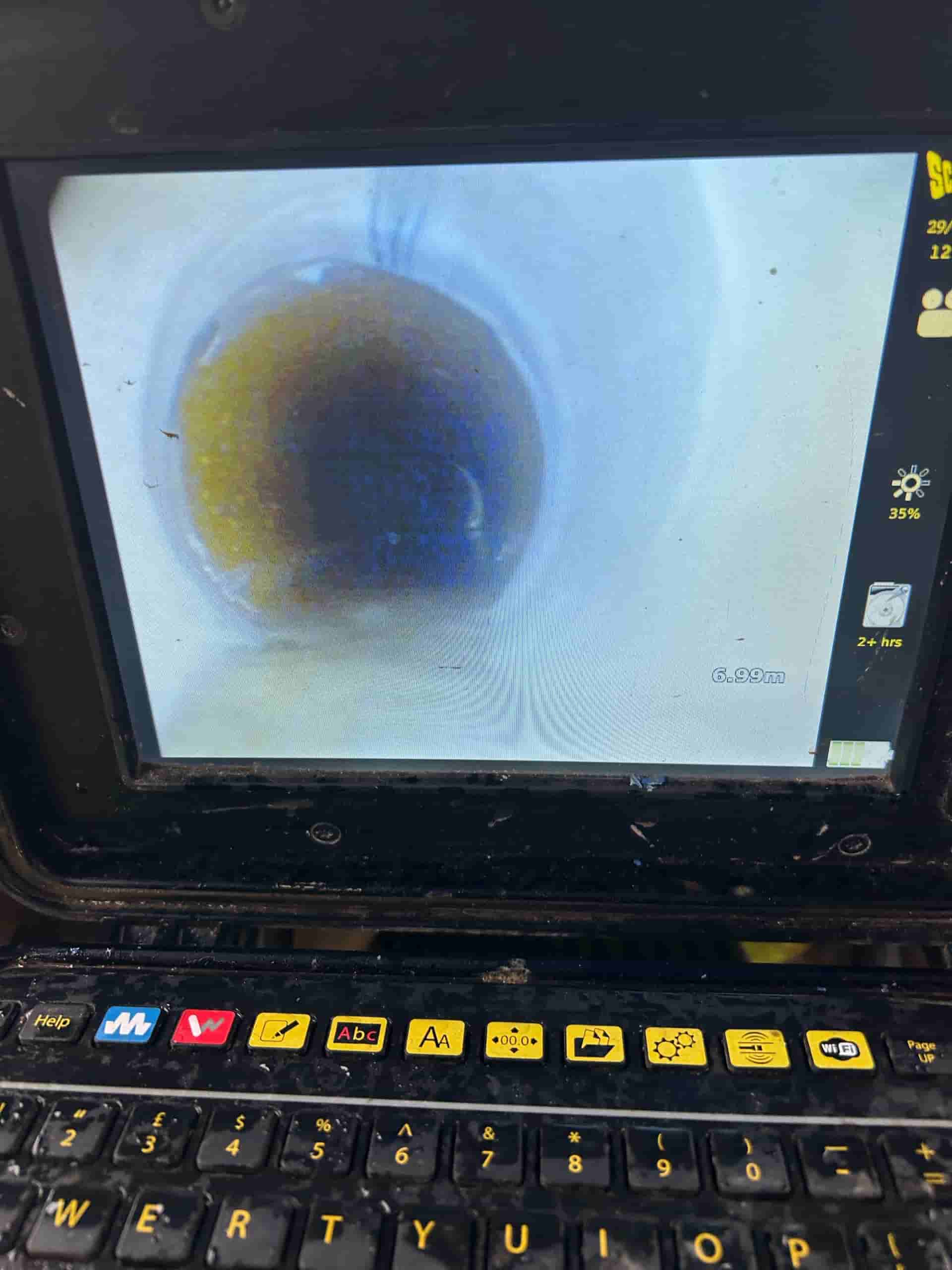 Close-up of a laptop screen showing a pipe inspection camera view inside a cylindrical pipe, revealing a partially blocked or dirty section. The computer's keyboard and interface buttons are visible below the screen.
