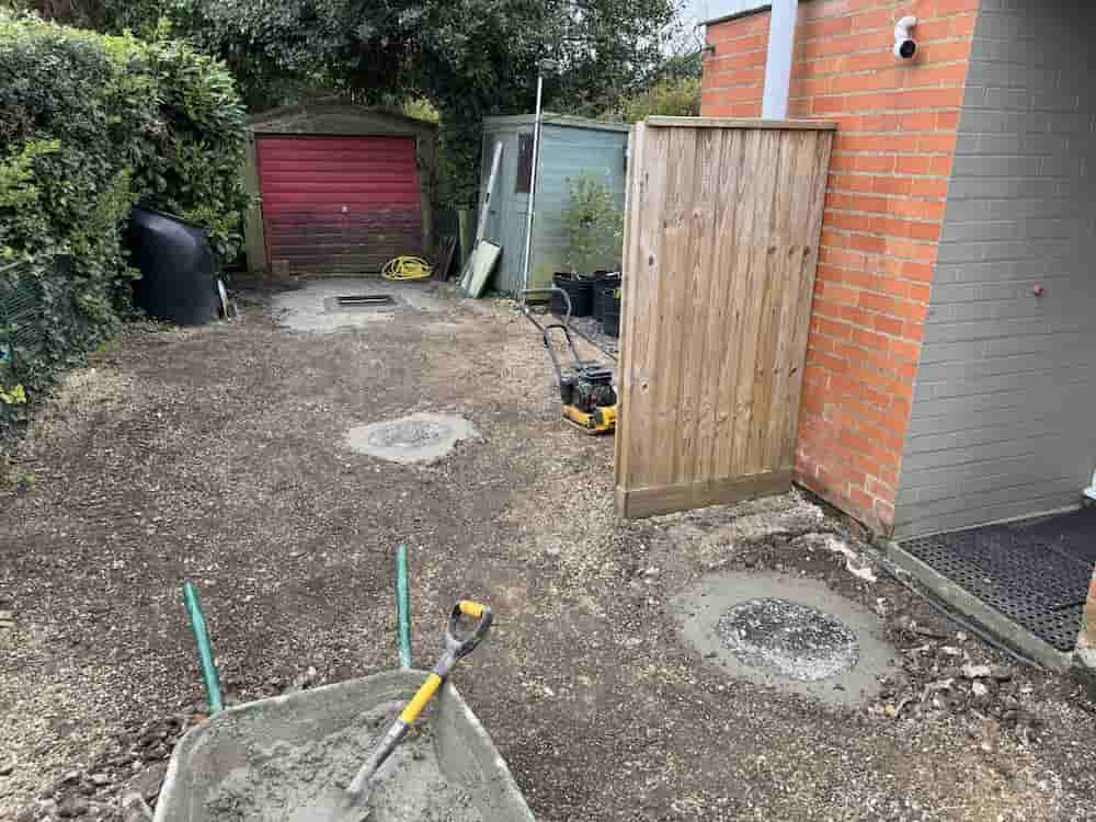 A partially cleared driveway with patches of wet concrete, garden tools, a wheelbarrow with a shovel, a red garage, and a wooden fence next to a brick house. Bushes and a shed are in the background.