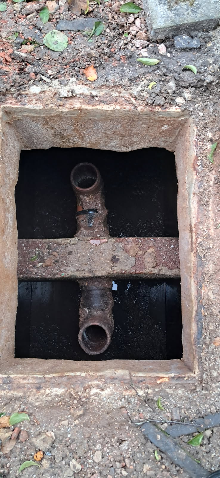 A rectangular open manhole reveals two rusty metal pipes running parallel, surrounded by dark soil and a concrete frame, with scattered leaves and debris around the opening.