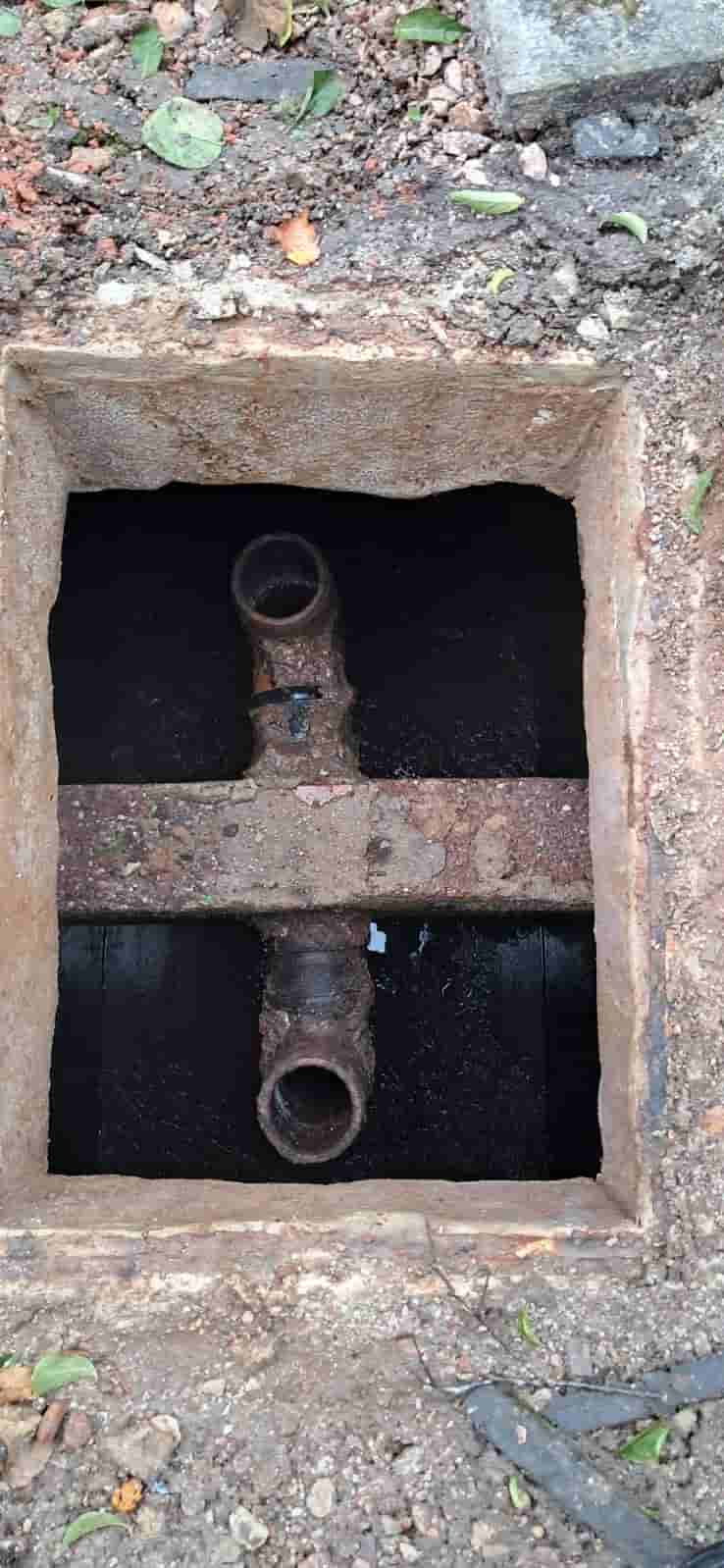 A rectangular concrete pit with two rusted metal pipes crossing each other inside, surrounded by dirt, rocks, and a few scattered leaves.