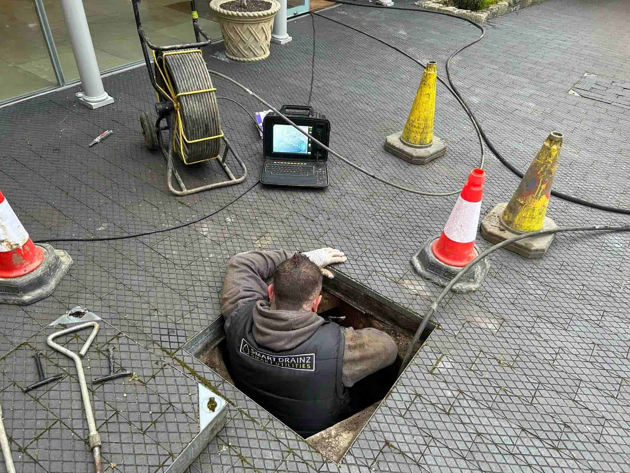 A worker in a vest is inside a maintenance hole, surrounded by orange and white traffic cones. Nearby, there is equipment, cables, and a laptop displaying a camera feed of the pipes.