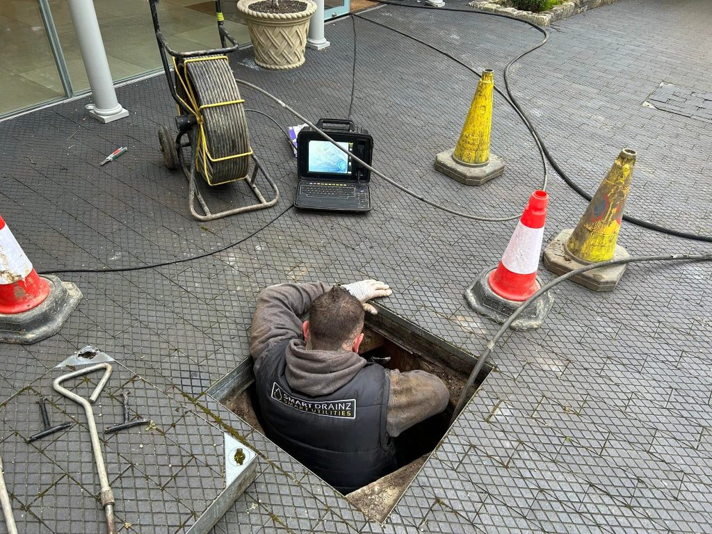 A worker wearing a "North Drains" vest is partially inside an open manhole, surrounded by traffic cones and drain inspection equipment. A laptop displays camera footage next to the worker on the tiled ground.