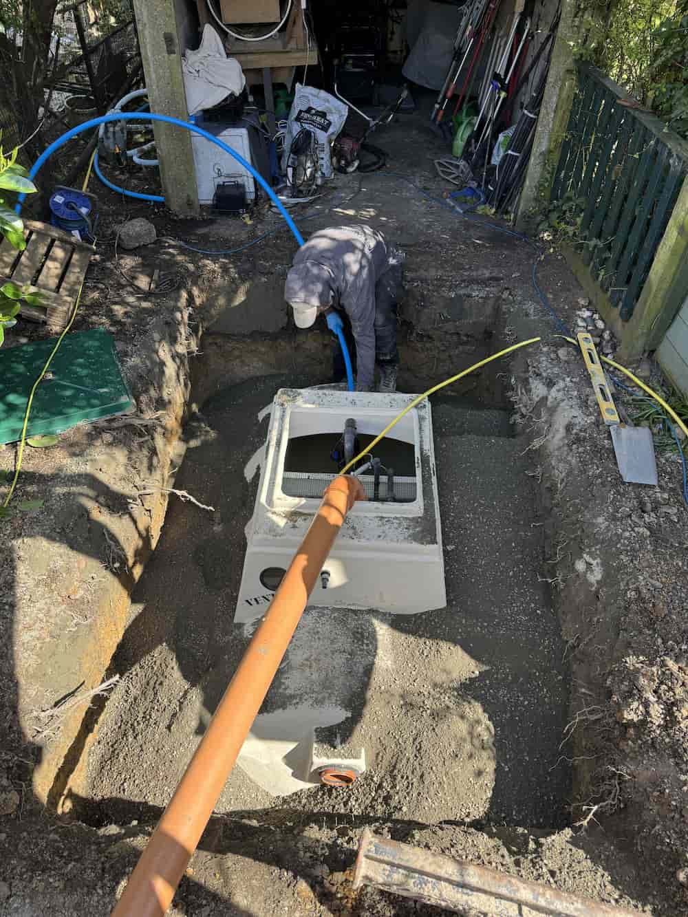 A person in work clothes installs a large concrete utility box or septic tank in a dug-out pit in a backyard, surrounded by tools, pipes, and a wheelbarrow near a shed or garage.