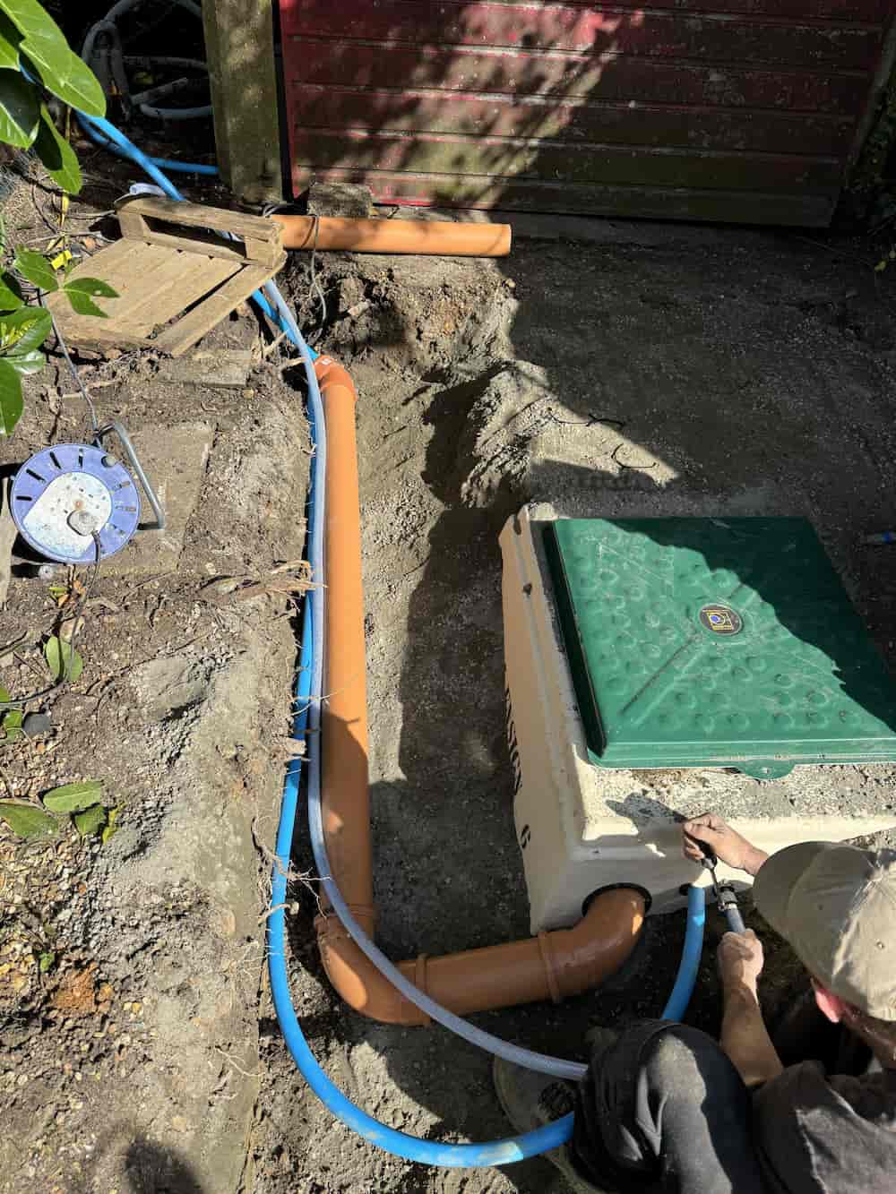 A person installs underground pipes and a water meter box in a dug trench near a building with a red wall. Blue and orange pipes are visible, along with tools and a cable reel on the ground.
