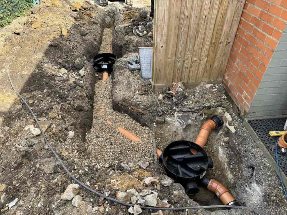 A trench dug alongside a house with exposed orange drainage pipes, gravel backfill, and two black inspection chamber covers, next to a brick wall and a wooden fence.